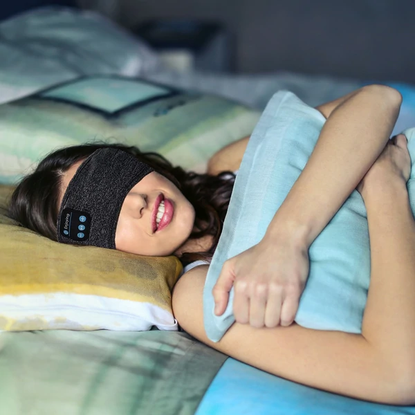 Gray Bluetooth sleep mask worn in bed, contoured band with flat built-in speakers